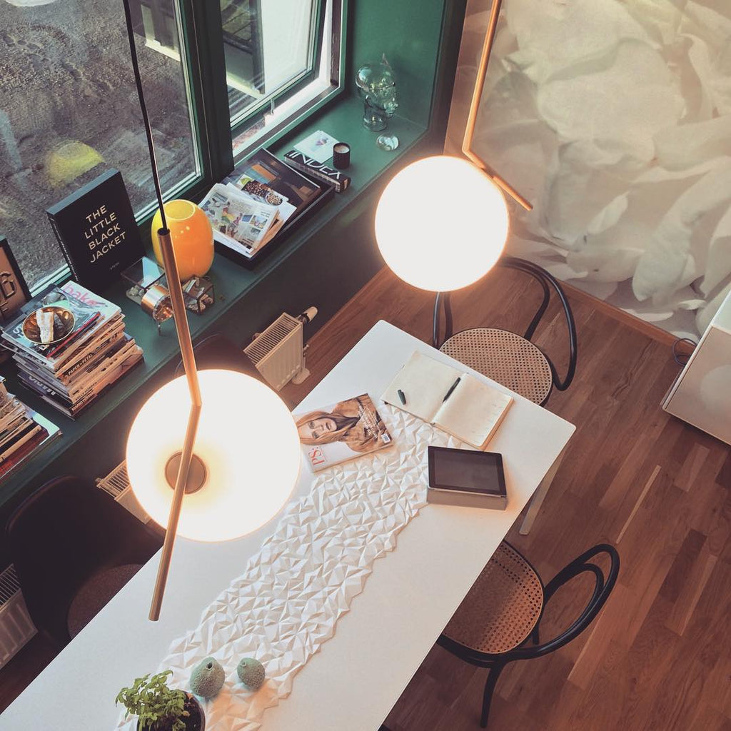A Clérachic pendant glows over a white table runner in a photograph taken from above, with a window ledge of magazines beyond.