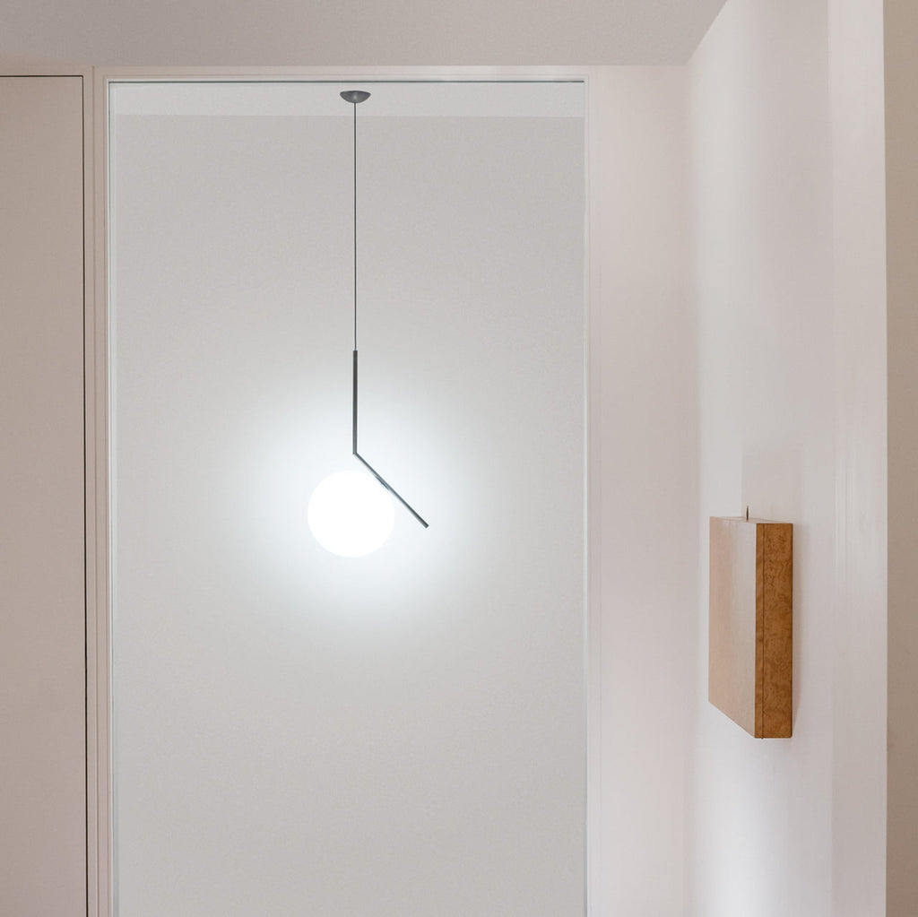 A Clérachic pendant with a slim dark stem hangs in a pale doorway, its white glass globe brightly lit beside a timber wall panel.