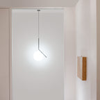 A Clérachic pendant with a slim dark stem hangs in a pale doorway, its white glass globe brightly lit beside a timber wall panel.