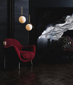 Two Clérachic globe pendants with a warm glow hang over a red patterned armchair in a dark, moody room with a dark floor.