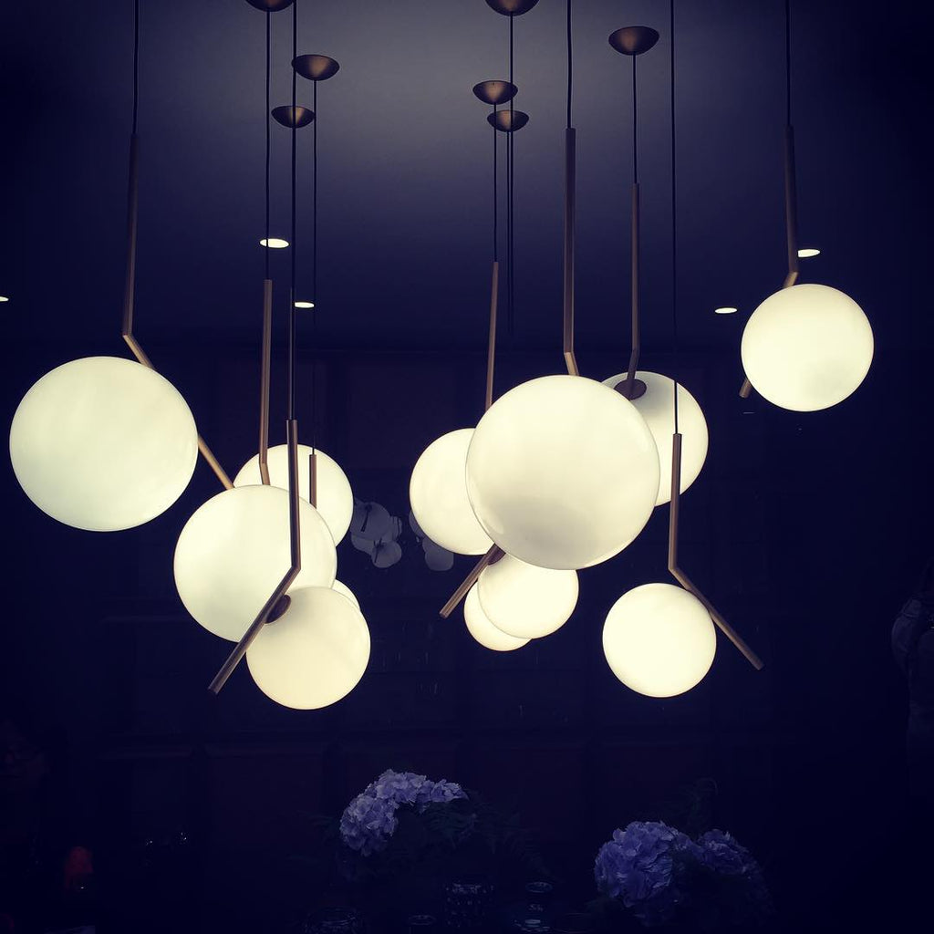 Many lit Clérachic globe pendants at varied heights fill a navy room, their brass stems angled over round white shades.