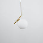 A Clérachic globe pendant on a pale grey backdrop, with a brass stem and a matt white glass ball hanging off-centre.