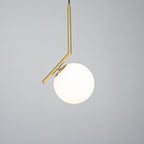 Lit Clérachic pendant on a mid-grey studio backdrop, with a bright white globe and a brass stem that bends above it.