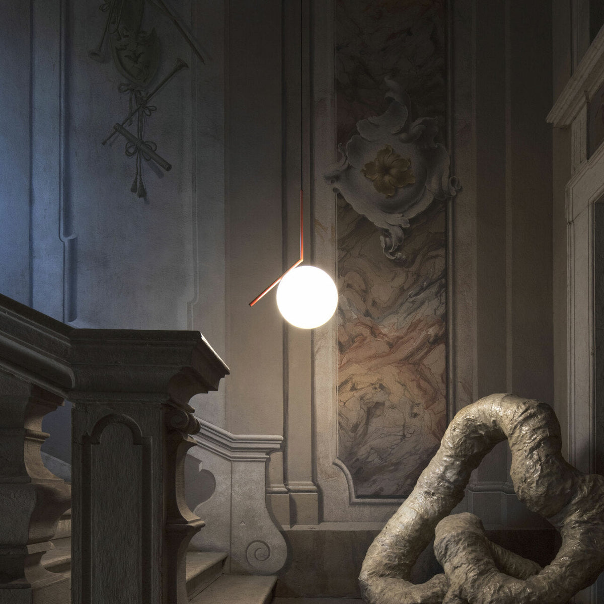 A Clérachic globe pendant hangs in a shadowy stairwell with a carved stone banister, a painted wall and a sculpture.