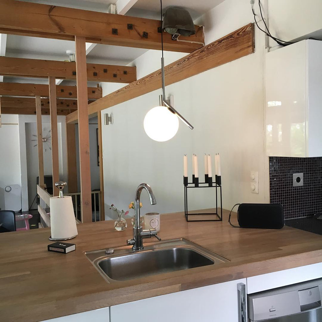 A Clérachic globe pendant hangs from a ceiling beam over a wooden kitchen counter, a steel sink and a black-tiled splashback.