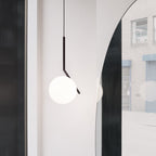 A Clérachic pendant with a black angled stem and a white glass globe hangs beside an arched mirror and a brick wall outside.