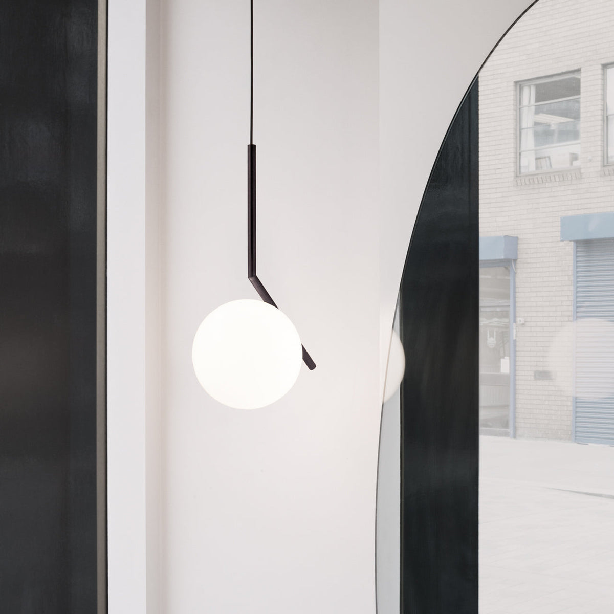 A Clérachic pendant with a black angled stem and a white glass globe hangs beside an arched mirror and a brick wall outside.
