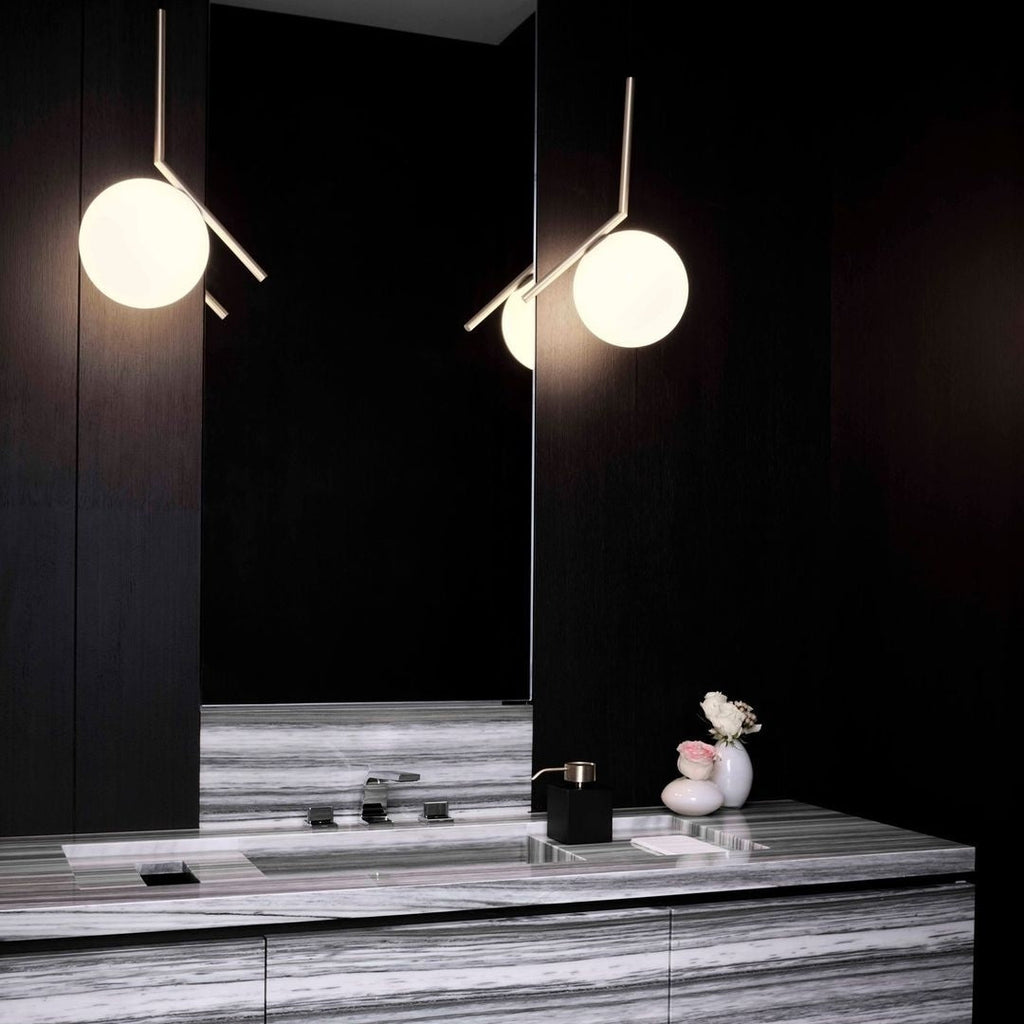 Two Clérachic globe pendants with brass stems glow in a black-panelled bathroom, one reflected above a striped marble vanity.