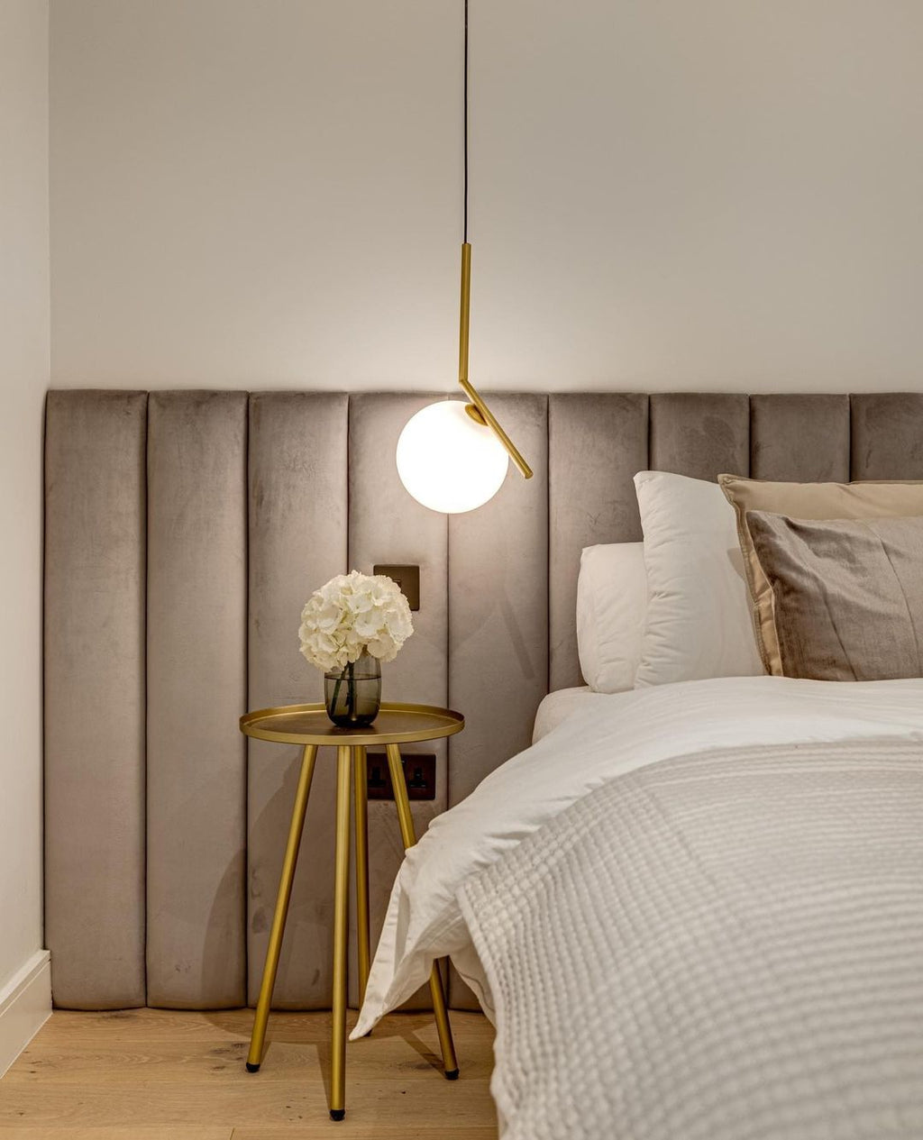 A Clérachic pendant with a brass stem and white globe hangs beside a velvet headboard, over a gold side table with white flowers.