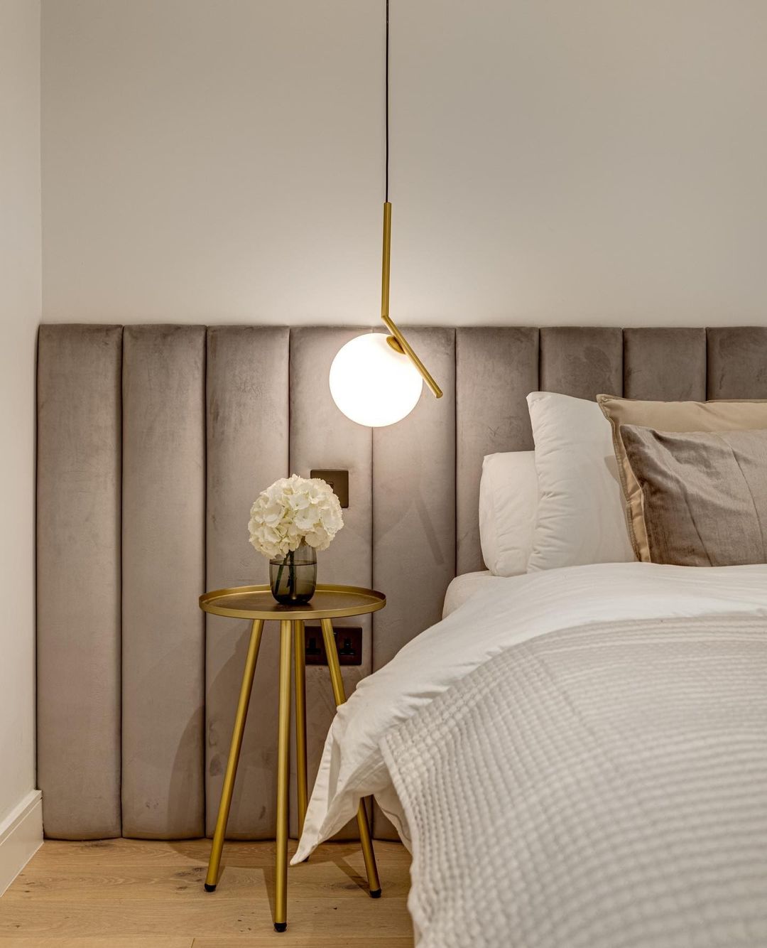 A Clérachic pendant with a brass stem and white globe hangs beside a velvet headboard, over a gold side table with white flowers.