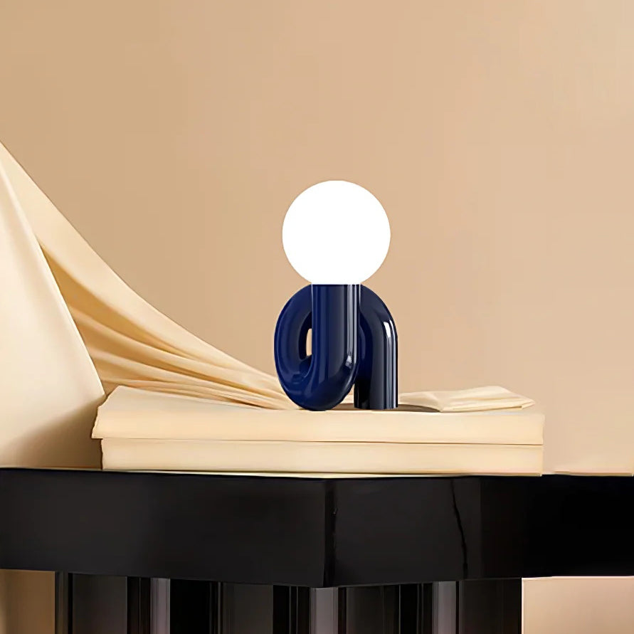 Blue Clérachic globe lamp on a cream book stack above a black table, with soft ivory fabric draped behind.