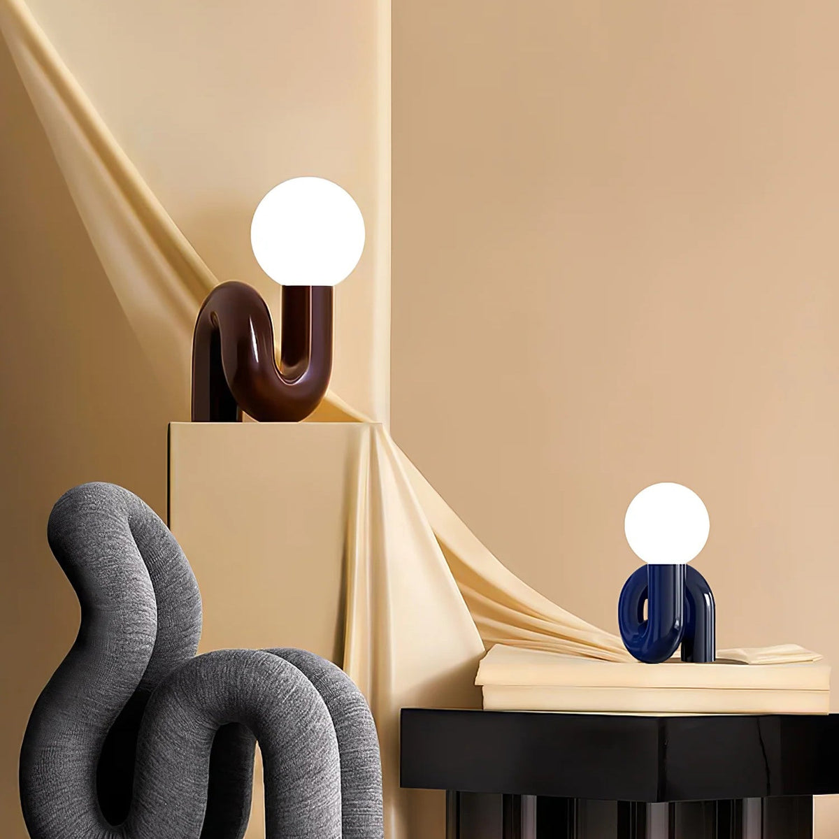 Dark red and blue Clérachic globe lamps on cream plinths against a warm beige wall, with a grey sculpture below.