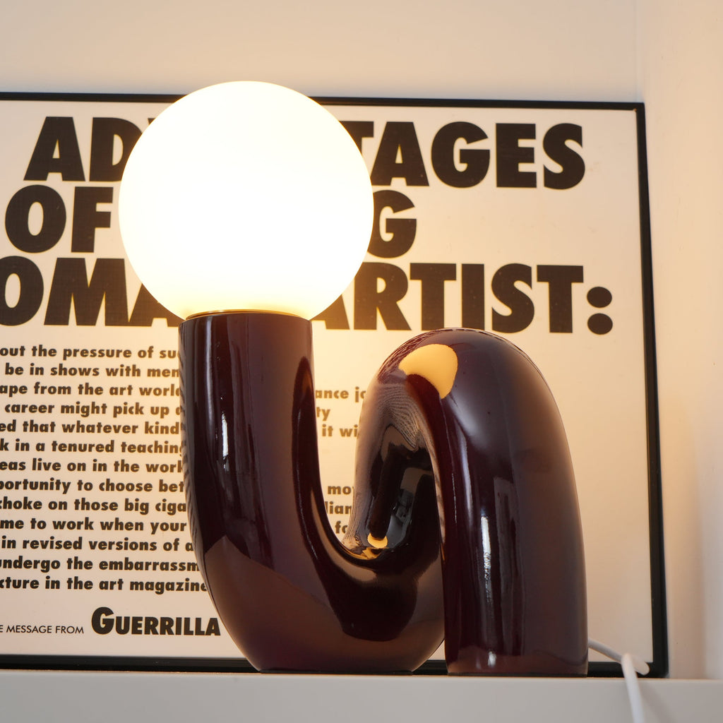 Lit dark red Clérachic table lamp with a warm glowing globe, its glossy tube base in front of a framed print.