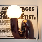 Lit dark red Clérachic table lamp with a warm glowing globe, its glossy tube base in front of a framed print.