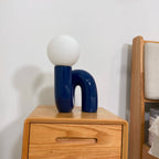 Blue Clérachic table lamp on a wooden bedside chest, its glossy U-shaped base beside a headboard.