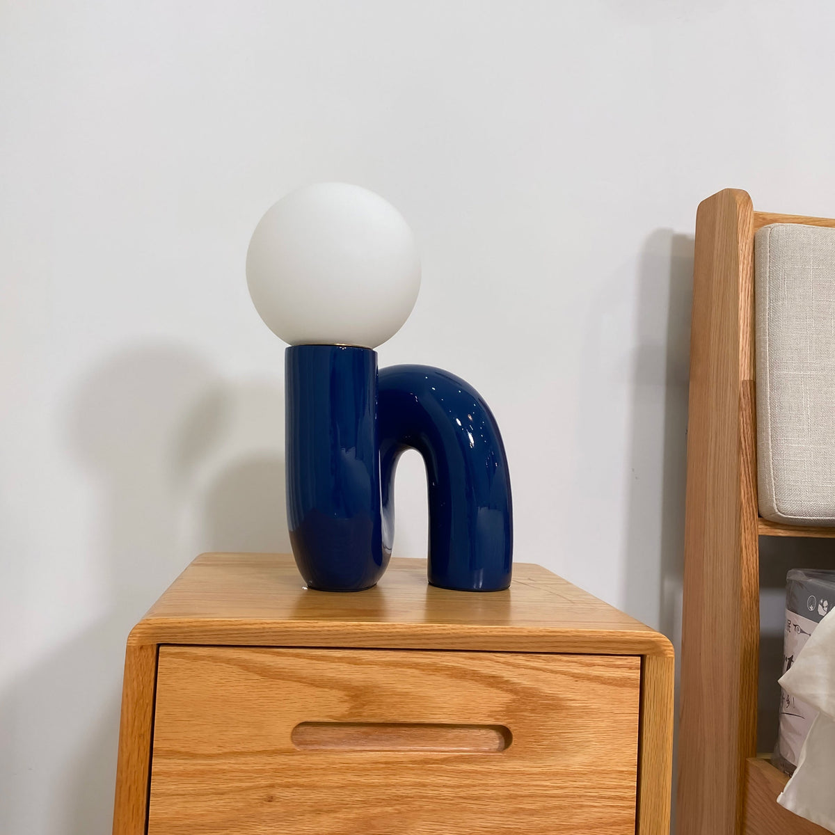 Blue Clérachic table lamp on a wooden bedside chest, its glossy U-shaped base beside a headboard.