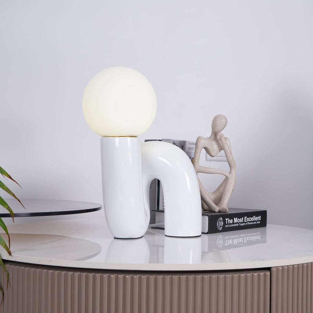 White Clérachic table lamp on a marble-topped cabinet, with a small seated figure and a book beside it.