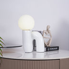 White Clérachic table lamp on a marble-topped cabinet, with a small seated figure and a book beside it.