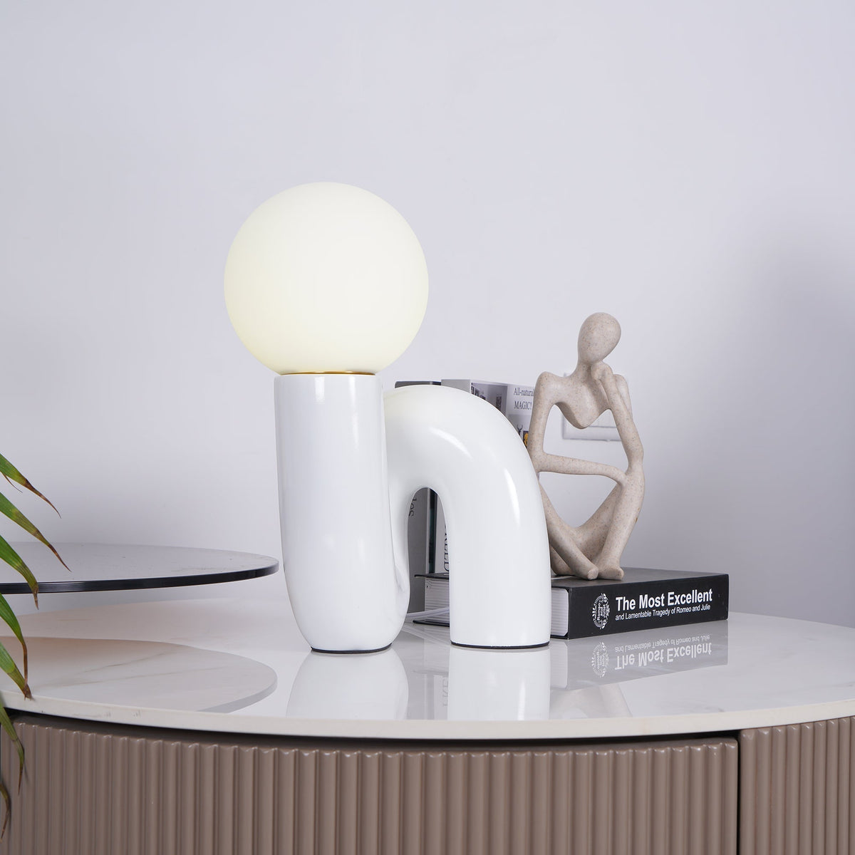 White Clérachic table lamp on a marble-topped cabinet, with a small seated figure and a book beside it.