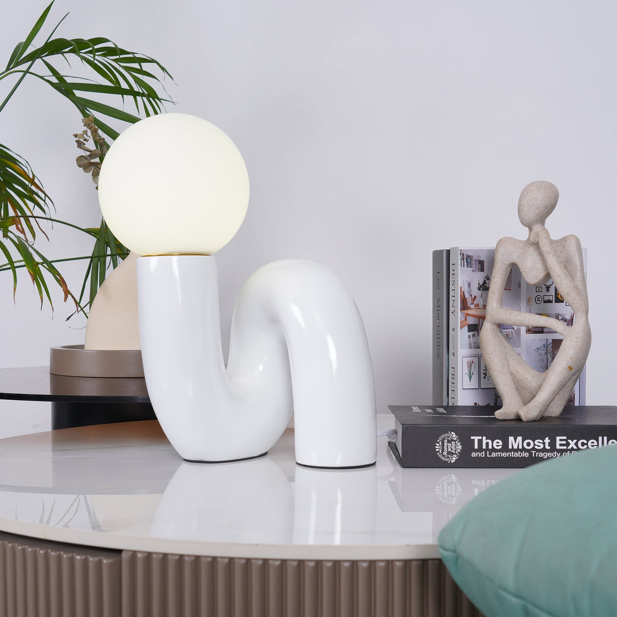 Glossy white Clérachic lamp on a pale marble top, with a stone figure and books, a green cushion at the edge.
