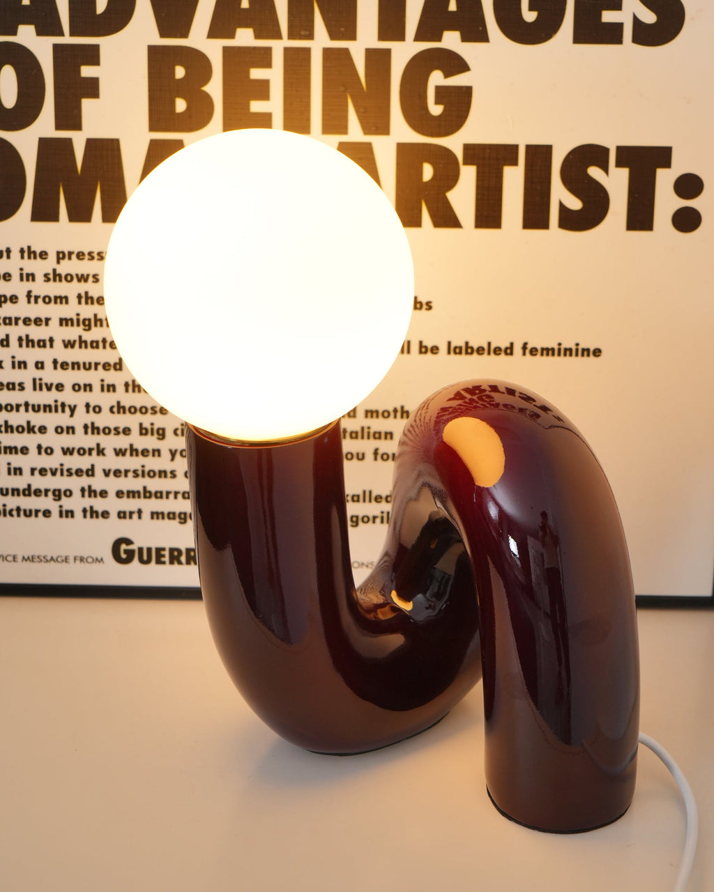Dark red Clérachic table lamp lit warm, its glossy looped base glowing from inside at the neck.