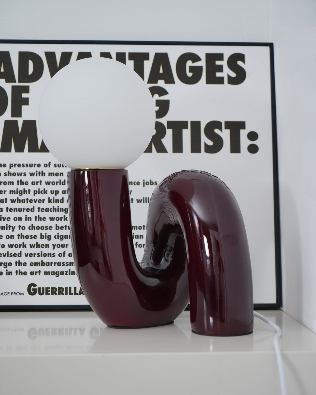 Dark red Clérachic table lamp unlit on a pale shelf, its glossy curved base reflected in the surface.