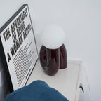 Dark red Clérachic table lamp seen from the side on a white shelf, with its cord trailing behind.