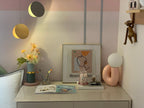 Small pink Clérachic lamp on a white desk with flowers, a framed print and hanging gold discs overhead.