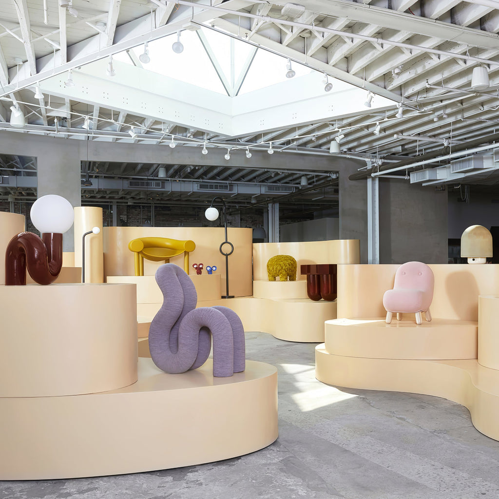 In a pale showroom with curved plinths, a dark red Clérachic globe lamp stands beside a lilac sculpture.