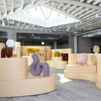 In a pale showroom with curved plinths, a dark red Clérachic globe lamp stands beside a lilac sculpture.