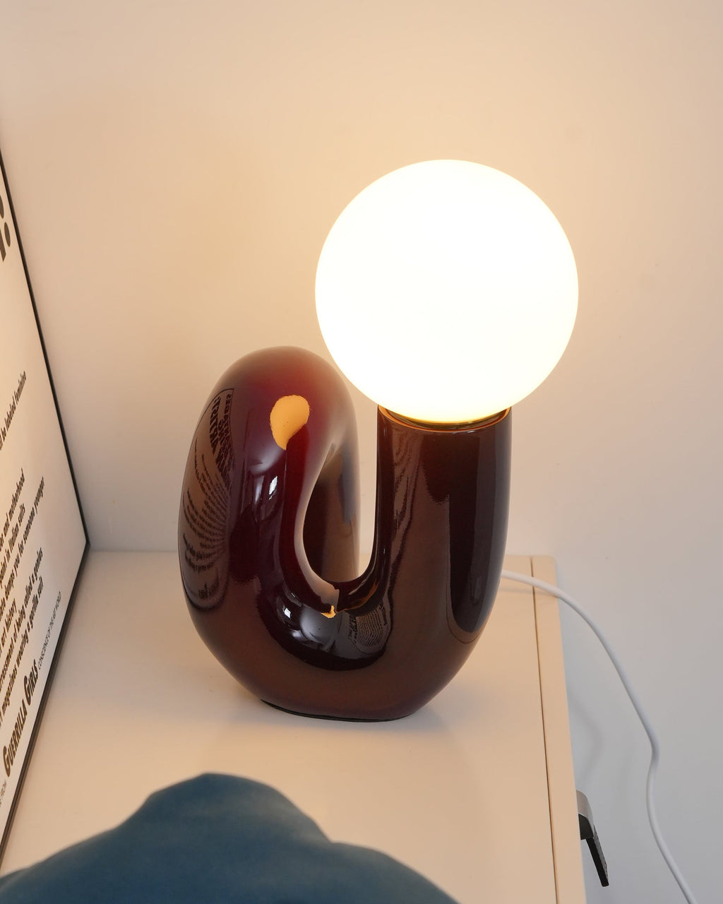 Dark red Clérachic table lamp lit on a white shelf, warm light glowing through the curve of its base.
