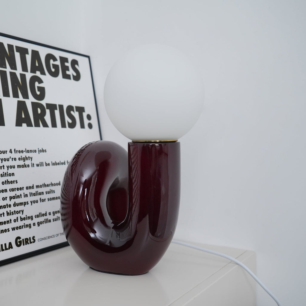 Glossy dark red Clérachic table lamp with an unlit globe, set on a white shelf against a white wall.