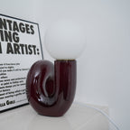 Glossy dark red Clérachic table lamp with an unlit globe, set on a white shelf against a white wall.