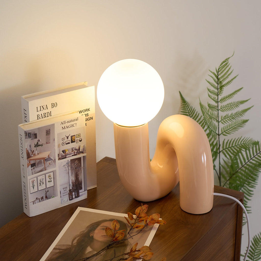 Lit peach Clérachic table lamp on a walnut chest, beside a book, a fern frond and a printed photo.