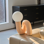 Peach Clérachic table lamp on the arm of a leather sofa, the globe near a window and a black cabinet.