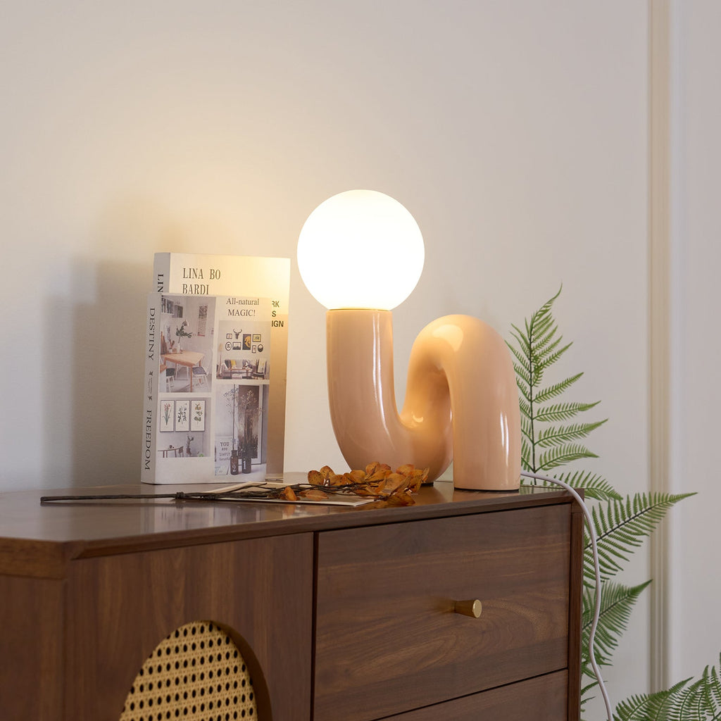 Peach Clérachic table lamp lit on a walnut cabinet with a cane panel, a fern and a book beside it.