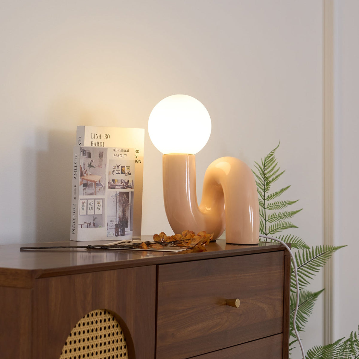 Peach Clérachic table lamp lit on a walnut cabinet with a cane panel, a fern and a book beside it.