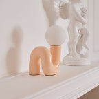 Peach Clérachic table lamp on a white mantel-style ledge, with a white cherub statue to its right.