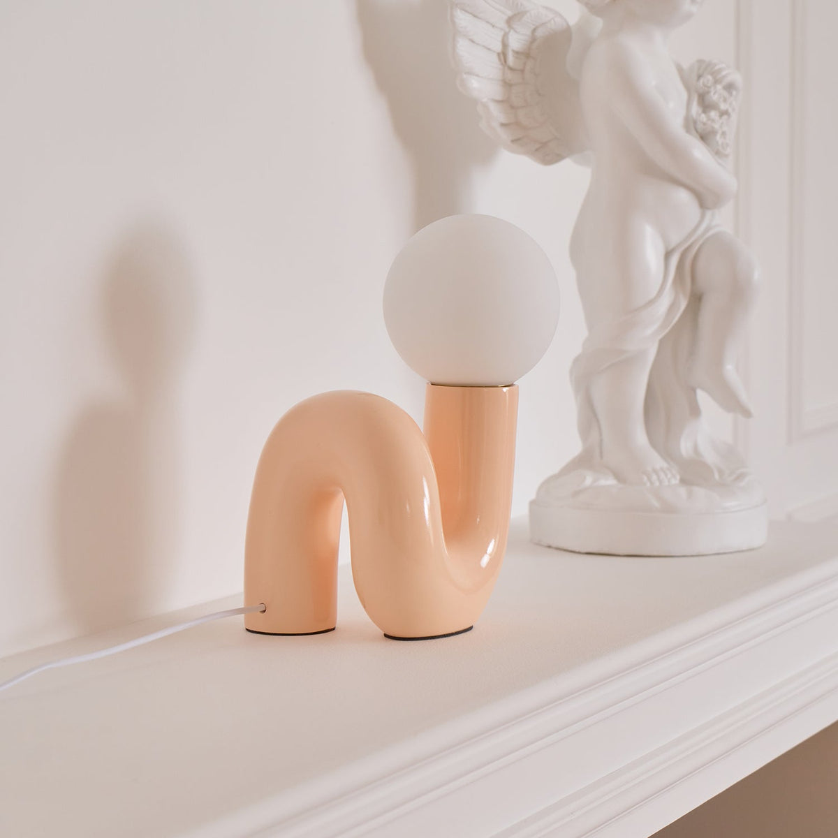 Peach Clérachic table lamp on a white mantel-style ledge, with a white cherub statue to its right.