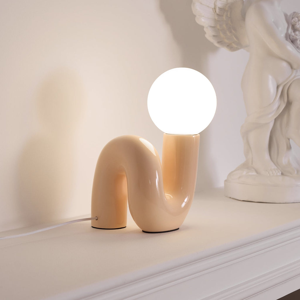 Lit peach Clérachic table lamp on a white ledge, its warm globe beside a cherub statue.