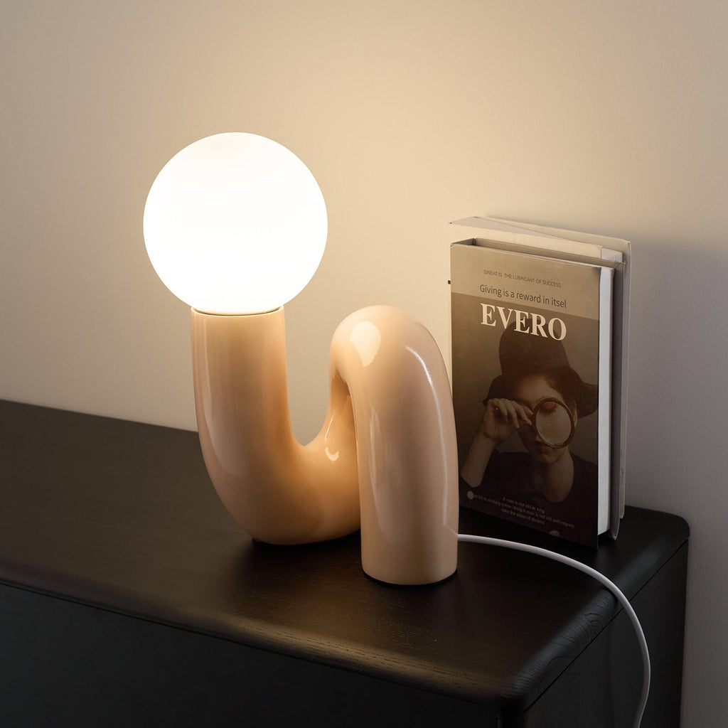 Peach Clérachic table lamp lit on a black cabinet, with a hardback book standing at its side.