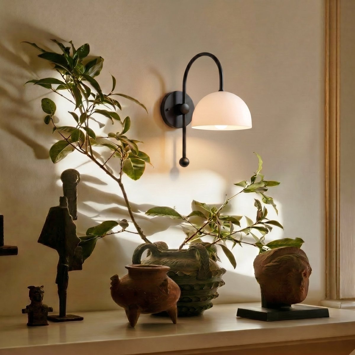 A black Clérachic wall lamp lights a mantel styled with small sculptures and potted greenery.