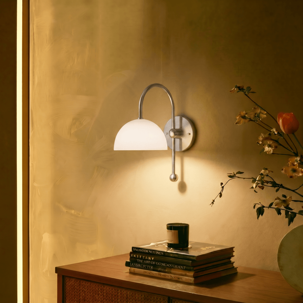 A chrome Clérachic wall lamp lights a console table stacked with books beside a candle and blossoms.