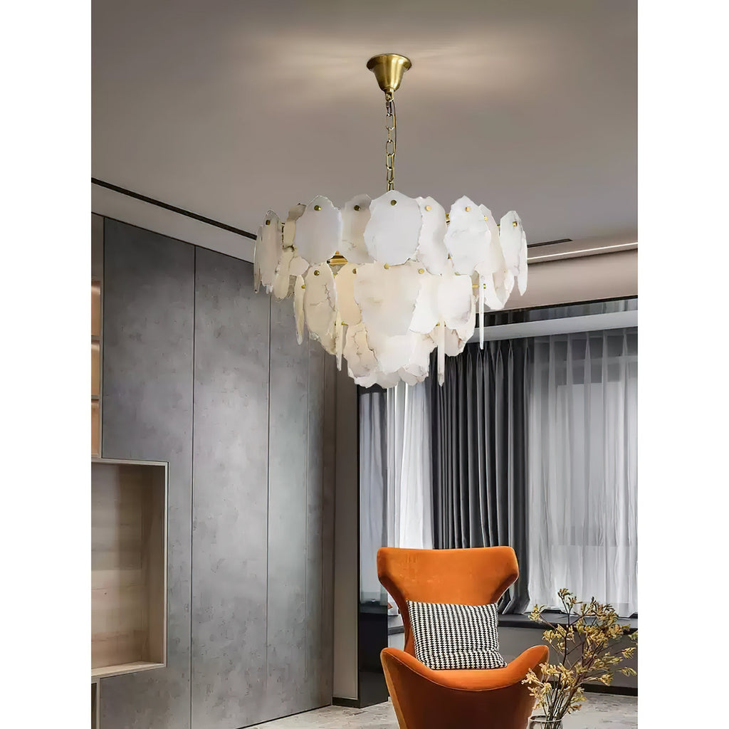 Clérachic Gloire chandelier against a concrete-panelled wall, glowing beside an orange velvet armchair and sheer curtains.
