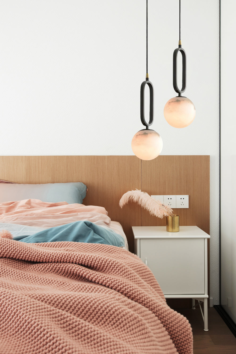 The Clérachic Gloruvyd pendants flank an oak headboard at uneven drops, warm stone light over pink and blue bedding.