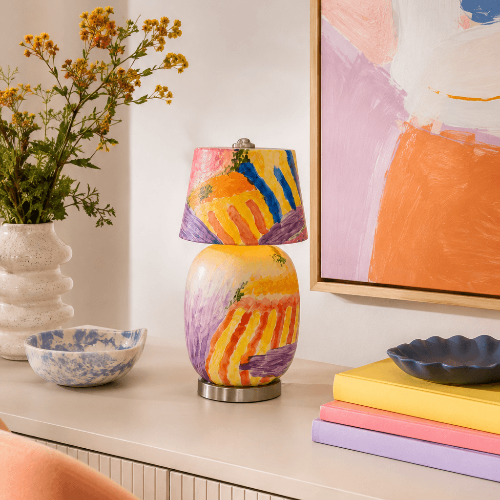 A table lamp painted in violet, orange, yellow and blue stands on a white sideboard with a pink-and-orange painting, a stack of pastel books and flowers, from Clérachic.