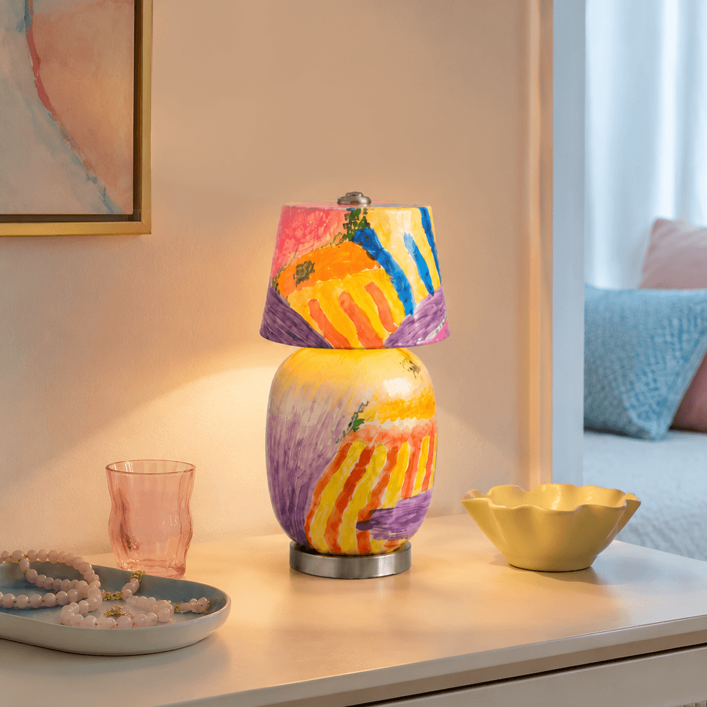 The painterly version of this Clérachic table lamp glows warm on a pale console beside a pink glass, a yellow scalloped bowl and a bead strand.
