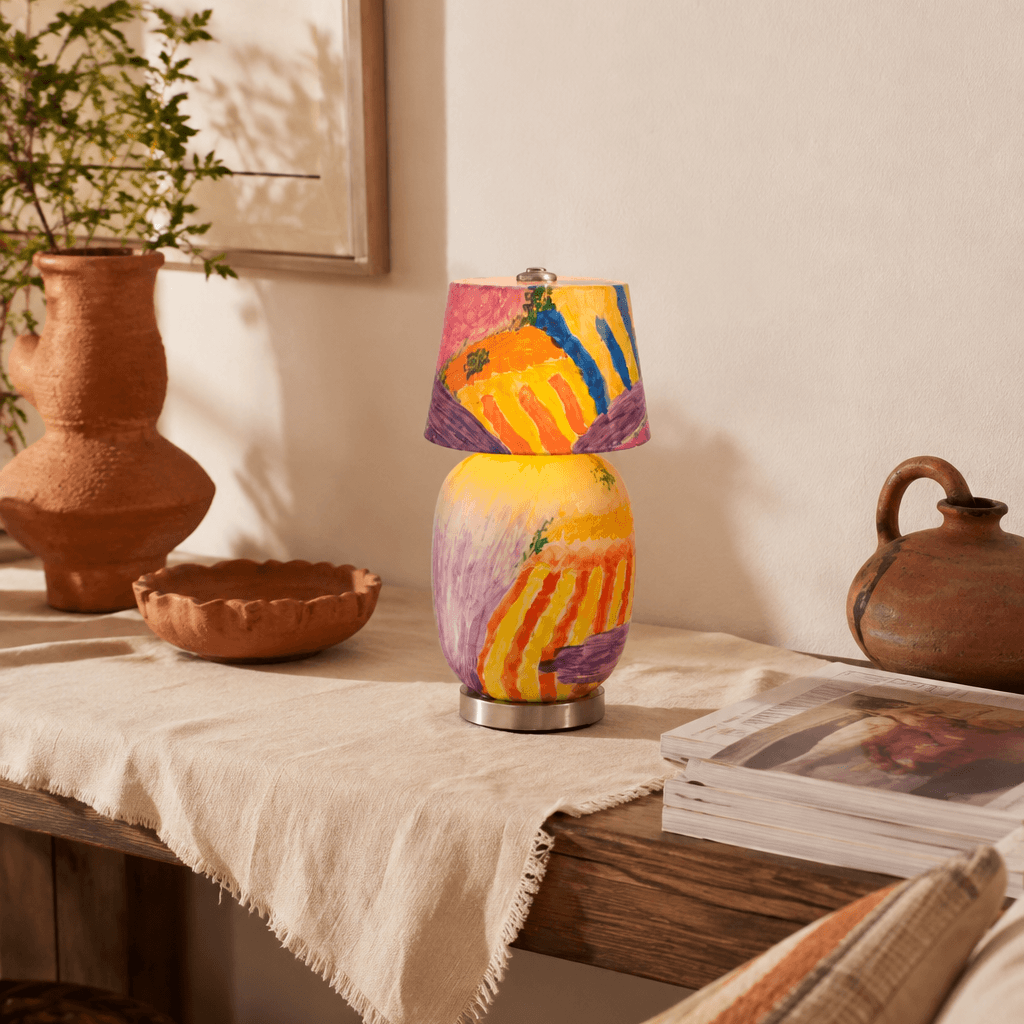 A small hand-painted lamp in yellow, orange and purple stands on a wooden table with terracotta pots and a magazine, a Clérachic table lamp.