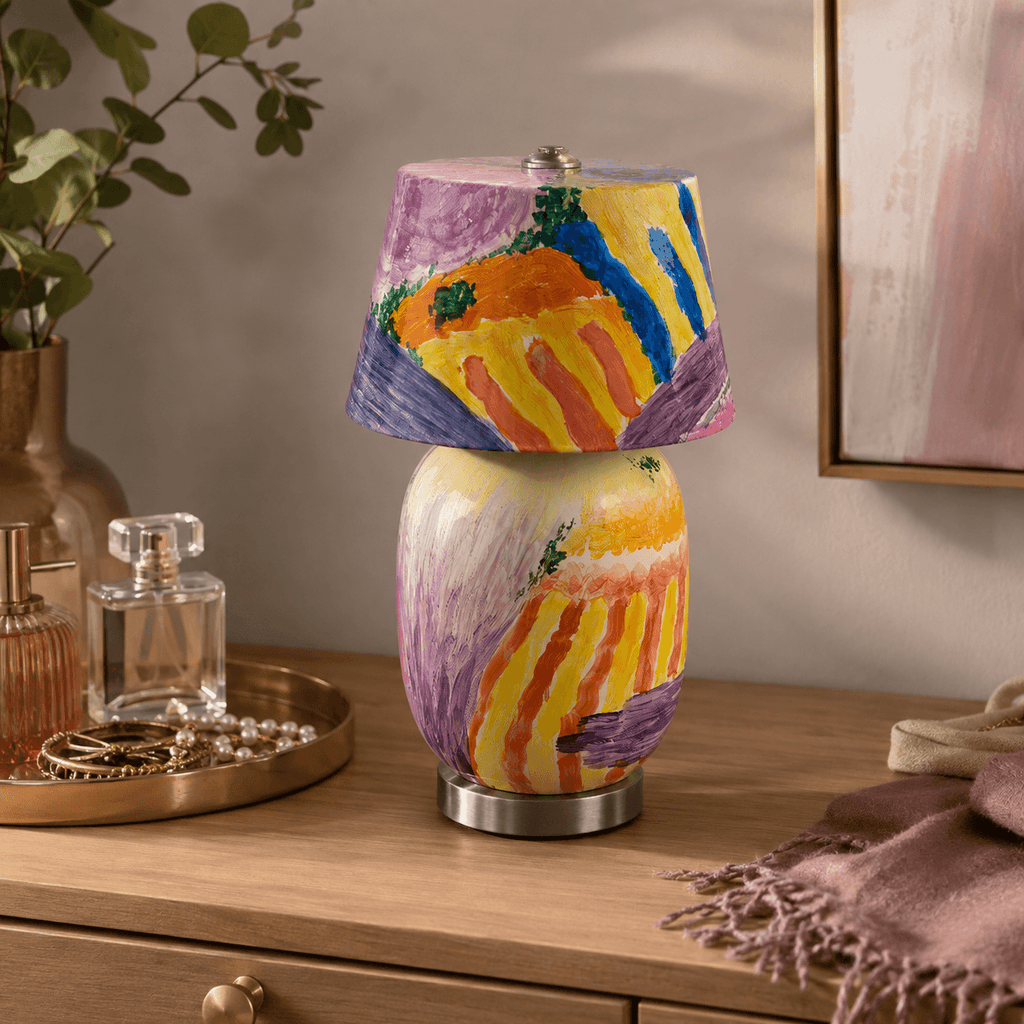 Close view of this Clérachic table lamp's glossy painted body in streaks of yellow, orange and violet, on a wooden dresser beside a perfume bottle and brass tray.