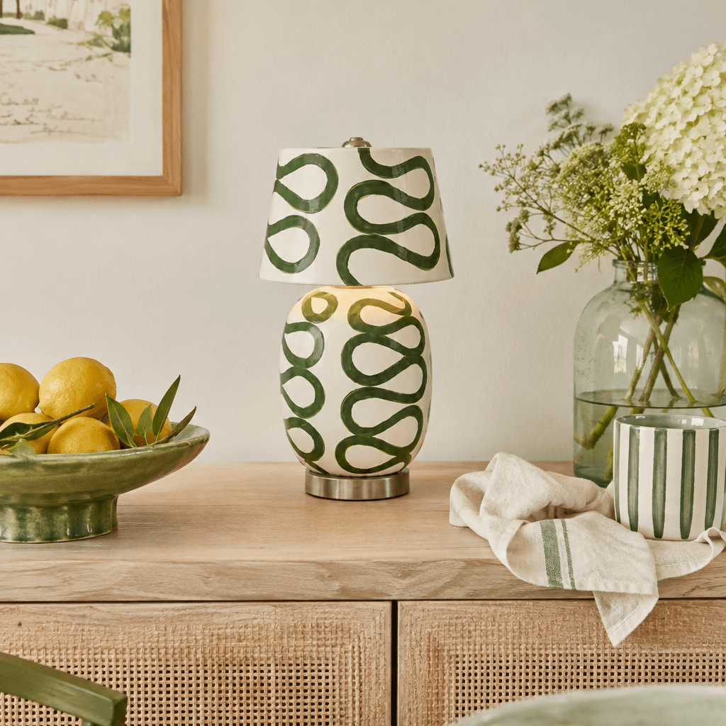 The green-looped version of this Clérachic table lamp stands on a cane-fronted sideboard beside a lemon bowl, a glass vase and white flowers.